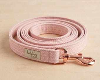 May include: A coiled light pink dog leash featuring a woven herringbone pattern. The leash includes a metallic rose gold swivel snap hook and a small rectangular fabric label with cursive text stitched onto the end of the strap.