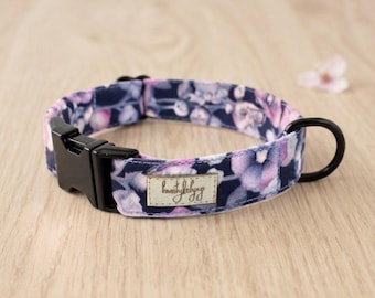 May include: A pet collar featuring a dark navy blue fabric with a floral pattern in shades of purple and pink. The collar includes a black plastic side-release buckle, a black metal D-ring, and a rectangular beige fabric label with cursive text.
