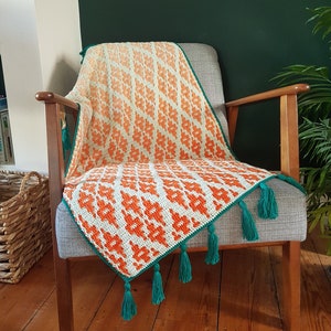 Op de afbeelding: Een gehaakt deken met een geometrisch patroon in oranje en wit, met een groene rand en kwastjes. Het deken is gedrapeerd over een houten fauteuil met een grijs kussen.