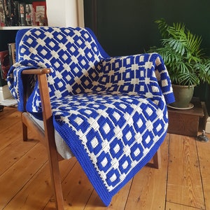 Peut inclure: Une couverture en crochet bleu et blanc avec un motif géométrique est drapée sur un fauteuil en bois. La couverture est faite avec un motif carré de carrés bleus et blancs.