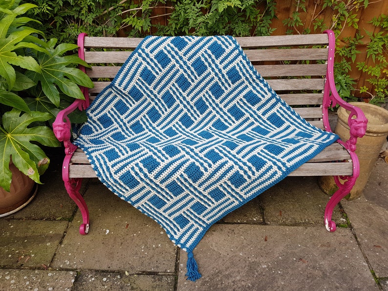 May include: A blue and white crocheted throw blanket with a geometric pattern, draped over a wooden bench with pink painted metal legs.