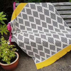 Puede incluir: Una manta de crochet blanco y negro con un borde amarillo, drapeada sobre un banco de madera rosa.