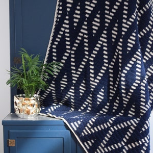 May include: A navy blue and white crocheted throw blanket with a geometric pattern. The blanket is draped over a blue cabinet with a plant in a gold pot.