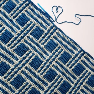 May include: A blue and white crocheted blanket with a geometric pattern. The blanket is partially finished and a blue crochet hook is lying on top of it.
