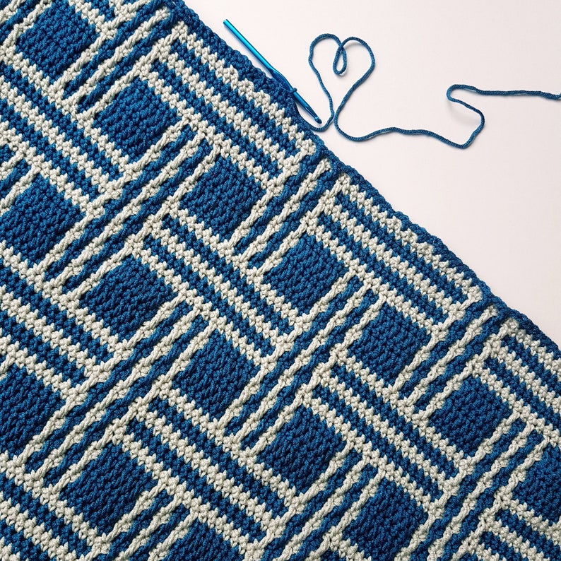 May include: A blue and white crocheted blanket with a geometric pattern. The blanket is partially finished and a blue crochet hook is lying on top of it.