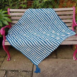 May include: A blue and white striped crocheted throw blanket with a tassel on a wooden bench.