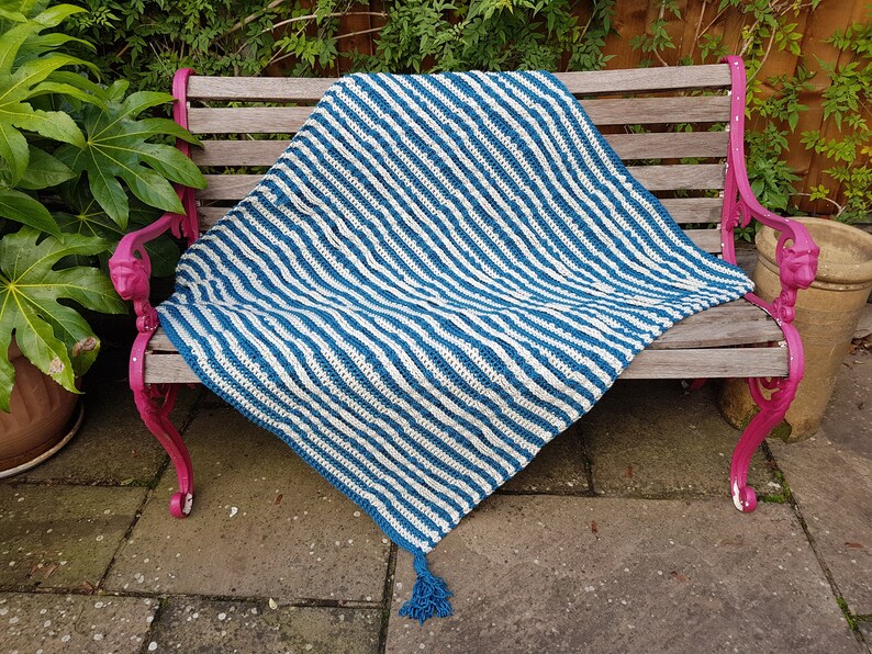 May include: A blue and white striped crocheted throw blanket with a tassel on a wooden bench.