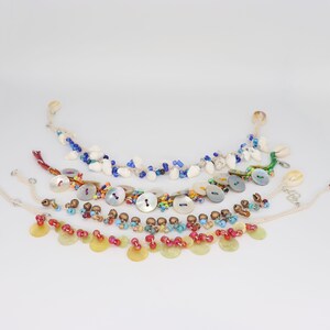 May include: Five colorful anklets with various beads, shells, and buttons. The anklets are made with white string and feature different colored beads, including red, blue, green, and brown. Some of the anklets have small shells and buttons.