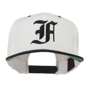 May include: Cream-colored baseball cap with a black brim and a black embroidered "FA" gothic-style letter design. The underside of the brim is green, and the cap has a black adjustable strap.