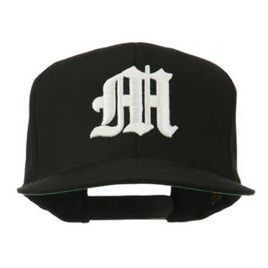 May include: Black baseball cap featuring a white embroidered letter "M" in a gothic font. The cap has a flat peak with a green underside and an adjustable strap at the back.