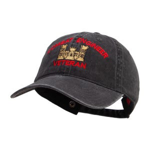 May include: A black, adjustable baseball cap with the words "COMBAT ENGINEER VETERAN" embroidered in red. The cap features a gold and red emblem of a castle. The brim is curved, and the cap has a metal clasp closure.