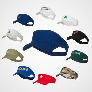 May include: Assortment of 12 different visor hats with various colors, designs, and text. The hats feature a variety of designs, including a shamrock, a pink ribbon, a Canadian flag, a US flag, and the text "Make America Great Again", "Police", "Irish", and "Vietnam".