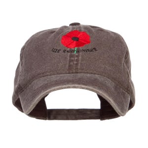 May include: A brown baseball cap with a red poppy flower embroidered on the front. The words "WE REMEMBER" are embroidered in black below the flower. The cap has a curved brim and an adjustable strap.