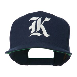 May include: A navy blue baseball cap with a flat peak and a white, stylised "K" embroidered on the front. The underside of the peak is green. A classic, casual accessory.