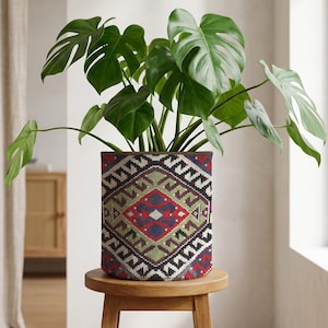 May include: A Monstera plant with large, green, split leaves in a patterned plant pot. The pot has a geometric design in red, green, and blue. The plant pot is on a small, round wooden stool. The pot's design is reminiscent of a rug.