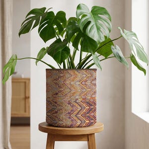May include: A Monstera plant with large, green leaves is displayed in a patterned plant pot. The pot has a design in shades of orange, purple, and gold. The plant pot is placed on a small, round wooden stool. The pot's design is reminiscent of a tapestry.