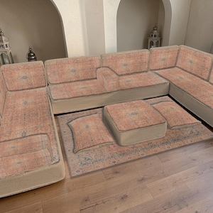 Op de afbeelding: Een grote hoekbank met bijpassende poef en vloerkleed. De bank en poef hebben een perzik-kleurige stof met patroon en een beige basis. Het vloerkleed heeft een vergelijkbaar patroon met blauwe accenten. De meubels staan op een parketvloer.