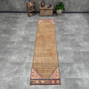 May include: A long, narrow runner rug featuring a central field of repeating geometric patterns in shades of tan and brown. The rug is framed by pink and dark blue borders with intricate floral and geometric motifs, finished with short fringe.