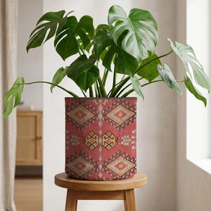 May include: A green Monstera plant in a cylindrical red planter with a geometric pattern. The planter sits on a small, round wooden stool. The plant's large leaves contrast with the patterned planter.