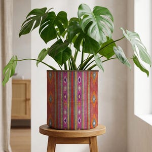 May include: A vibrant, patterned plant pot with a colorful design of vertical stripes and diamond shapes. The pot holds a lush, green Monstera plant, and sits on a small, round wooden stool. The pot's design includes red, orange, purple, and yellow hues.