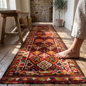 May include: A long, rectangular runner rug featuring a repeating geometric pattern in shades of deep red, orange, yellow, and black. The rug has a central row of diamond-shaped motifs bordered by intricate triangular and zigzag designs along the edges.