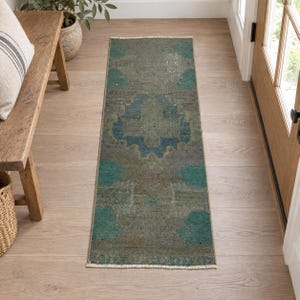 May include: A rectangular runner rug featuring a faded, distressed pattern in shades of sage green, teal, and muted blue. The rug has a central medallion design with geometric motifs and short, light-colored fringe along the two shorter ends.