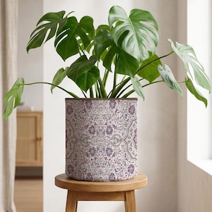 May include: A potted Monstera plant with large, green, split leaves. The plant is in a cylindrical pot with a floral pattern in shades of purple, gray, and white. The pot sits on a small, round wooden stool.