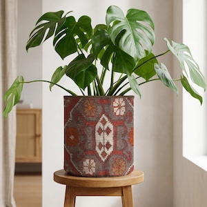 May include: A Monstera plant with large, green, split leaves in a patterned pot. The pot, with a design in red, brown, and white, sits on a small, round wooden stool. The plant pot is cylindrical in shape.