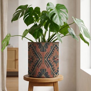 Może przedstawiać: Roślina Monstera w doniczce z dużymi, zielonymi, podzielonymi liśćmi. Cylindryczna donica ma wzór w odcieniach czerwieni, niebieskiego i brązu. Donica stoi na małym, okrągłym, drewnianym stołku. Średnica donicy to około 30 cm.