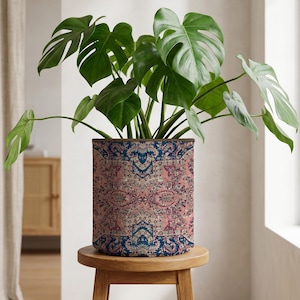 May include: A houseplant with large, green leaves in a patterned pot. The pot, with a design in pink, blue, and cream, sits on a small wooden stool. The pot's design resembles a traditional rug pattern.