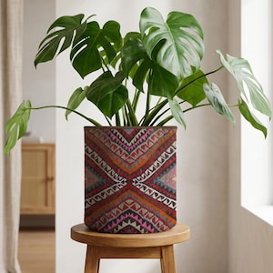 Op de afbeelding: Een grote groene Monstera-plant in een decoratieve plantenbak met een geometrisch patroon in rood, oranje en grijs. De plantenbak staat op een kleine, ronde houten kruk. De plant heeft grote, gespleten bladeren.