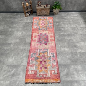 Puede incluir: Una alfombra pasillera larga y rectangular con un color base rojo vibrante y patrones geométricos repetitivos en tonos naranja, rosa, gris y blanco. La alfombra tiene un pelo texturizado y está rematada con flecos oscuros en los extremos.