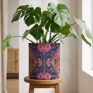 May include: A houseplant with large green leaves in a patterned cylindrical pot. The pot features a design in blue, pink, and brown. It is placed on a small, round wooden stool. The background shows a neutral-toned room with a window and a cabinet.