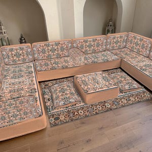 May include: A sectional sofa with a patterned fabric in shades of peach, green, and cream. The sofa is arranged on a matching area rug, with a matching ottoman and throw pillows. The sofa has a low profile and a modern design.