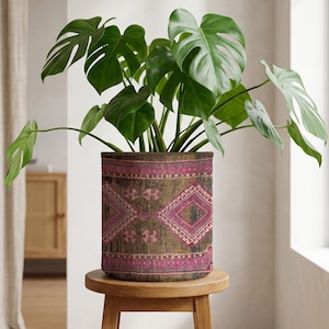Puede incluir: Una planta de Monstera en maceta con hojas verdes vibrantes en una maceta cilíndrica. La maceta presenta un patrón geométrico marrón y rosa, agregando un toque bohemio. La maceta se coloca sobre un pequeño taburete redondo de madera.