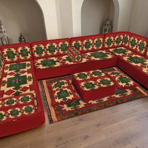 Puede incluir: Un sofá seccional rojo con cojines estampados y un juego de otomanas a juego. Los cojines presentan un diseño geométrico en verde, beige y rojo. Una alfombra coordinada con un patrón similar completa el conjunto de sala.