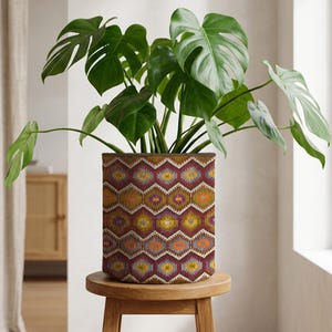 May include: A Monstera plant with large, green, split leaves in a patterned cylindrical pot. The pot features a geometric design in red, orange, yellow, and blue. It rests on a small, round wooden stool, creating a decorative display.