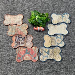 Puede incluir: Ocho posavasos de tela con forma de hueso que presentan varios patrones intrincados en tonos rojo, azul, beige y marrón. Una pequeña planta de hojas verdes y una figura en miniatura de un pez payaso naranja, blanco y negro están en el centro.