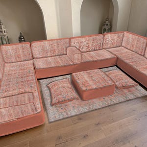 Puede incluir: Un gran sofá seccional de color coral con una tela estampada. El sofá está dispuesto en forma de L e incluye una otomana a juego y dos cojines. El sofá se encuentra sobre una alfombra estampada, creando un espacio habitable acogedor.