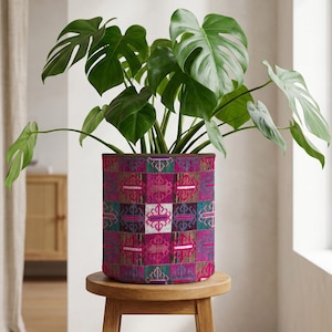May include: A green-leafed plant in a patterned cylindrical planter. The planter has a patchwork design in pink, purple, teal, and white. It sits on a small, round wooden stool. The plant pot is approximately 30 cm in diameter.