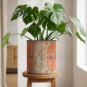 May include: A Monstera plant with large, green leaves in a cylindrical plant pot. The pot has a vintage design in orange, beige, and brown. The plant pot is placed on a small, round wooden stool. The pot's design is reminiscent of a Persian rug.