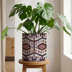 Op de afbeelding: Een cilindrische plantenpot met een geometrisch patroon in rood, blauw, oranje en groen. De pot bevat een grote groene plant met brede bladeren. De pot staat op een kleine, ronde houten kruk.