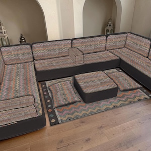 Puede incluir: Un sofá seccional con una tela estampada en tonos marrones, naranjas y verdes. El sofá tiene una base gris oscuro e incluye una otomana y una alfombra a juego. La alfombra y la otomana tienen la misma tela estampada que el sofá.