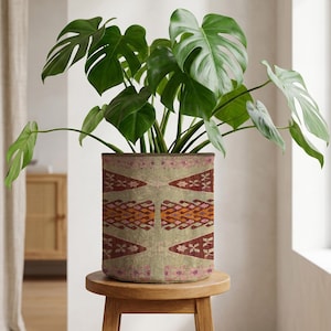 Peut inclure: Un grand Monstera aux feuilles vertes dans un pot de fleurs beige à motifs géométriques rouges, oranges et roses. Le pot est posé sur un petit tabouret rond en bois. L'arrière-plan comprend un mur de ton neutre et une armoire en bois.