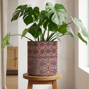 May include: A Monstera plant with large, green leaves in a cylindrical planter. The planter has a geometric pattern in red, orange, and white. The plant pot is on a small, round wooden stool. The pot's pattern is a repeating design.