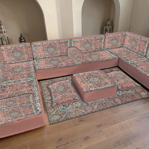 May include: A sectional sofa with a matching rug and ottoman set. The furniture features a pink base with a patterned fabric in shades of pink, blue, and cream. The set is arranged on a wooden floor, creating a cohesive living space.