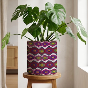 May include: A Monstera plant with large, green leaves in a cylindrical plant pot. The pot has a geometric pattern in purple, red, yellow, and white. The plant pot sits on a small, round wooden stool. The pot's pattern is reminiscent of a tribal design.