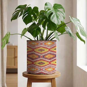 Puede incluir: Una planta de Monstera en maceta con grandes hojas verdes en una maceta estampada. La maceta presenta un diseño geométrico en rojo, naranja, amarillo y azul. La maceta está colocada sobre un pequeño taburete redondo de madera. El diámetro de la maceta es de unos 30 cm.