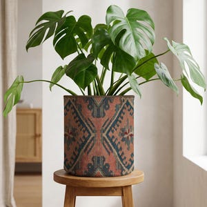 Puede incluir: Una planta de Monstera con hojas grandes, verdes y divididas en una maceta cilíndrica. La maceta tiene un diseño estampado en tonos rojos, azules y marrones. La maceta está sobre un pequeño taburete redondo de madera. La maceta mide unos 30 cm de diámetro.
