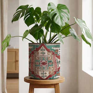 May include: A Monstera plant with large, green, split leaves in a cylindrical planter. The planter has a red, teal, and beige geometric pattern and sits on a small, round, wooden stool. The plant pot is approximately 25 cm in diameter.